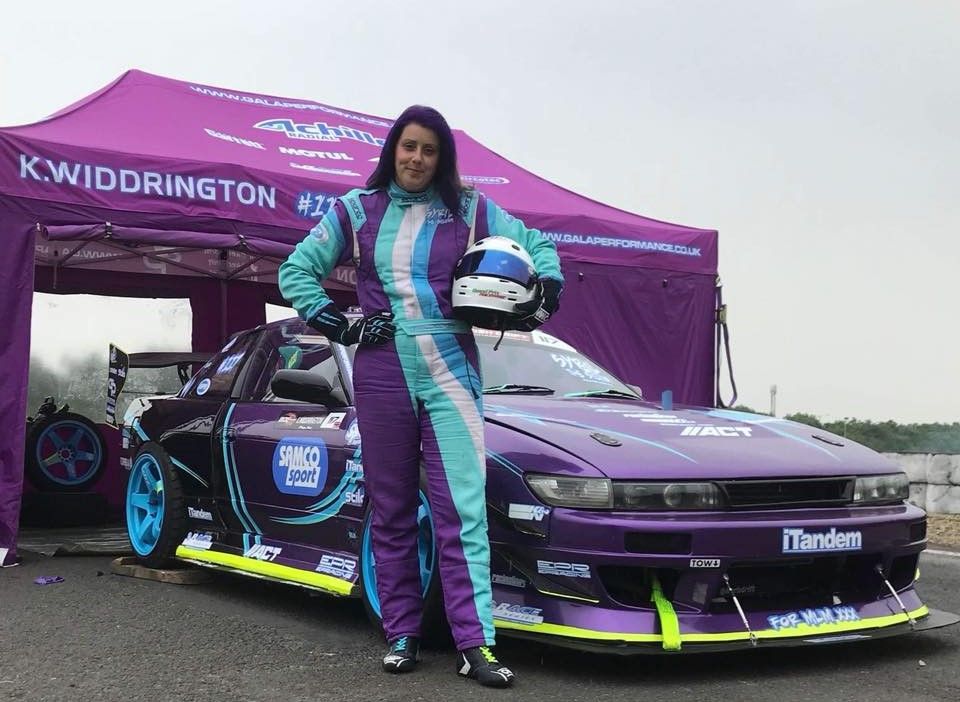 A female drift driver stands with her purple car beside a purple branded gazebo