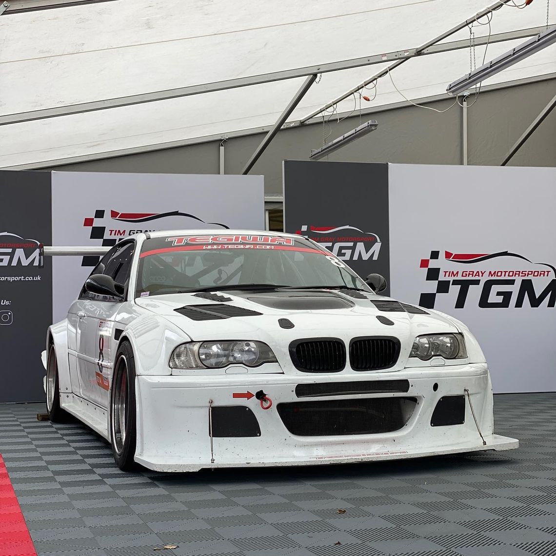 A racing car in front of some fabric pit walling with the branding for Tim Grey Motorsport